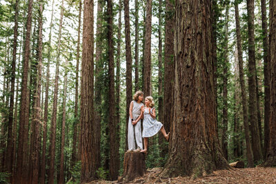 sisters standing on tree stump in the the woods of Joaquin Miller Park of Oakland interacting with each other