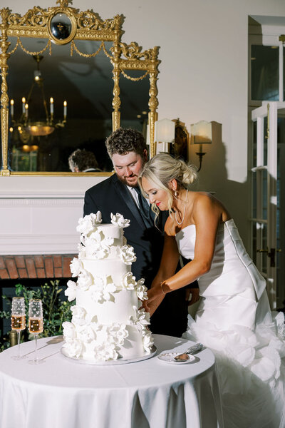 Kailea and Zach cut their wedding cake at Boxwood Estate, smiling as they share the moment. The elegant cake, decorated with delicate flowers and intricate designs, sits on a beautifully set table. Kailea wears a stunning white wedding gown, and Zach is dressed in a sharp suit. The warm lighting and sophisticated decor of the estate create a romantic and festive atmosphere, as the couple celebrates their special day.