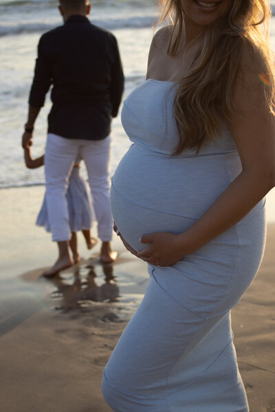 Mother holding her newborn while her two older children play at the water's edge on the beach