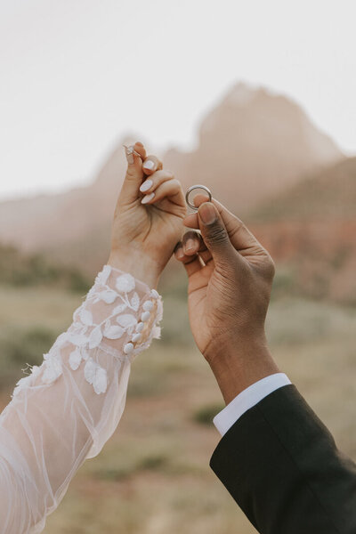 A close up shoot of a bride and groom holding their wedding rings, cropped so you only see from the elbow to their hands. Behind them, the blurred landscape of Torrey, Utah.