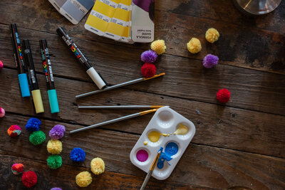 The enchanting world of art at Soleil Vida Studio. On a wooden table, discover a palette of creativity with colored paints, markers, brushes, and an array of vibrant pom-poms