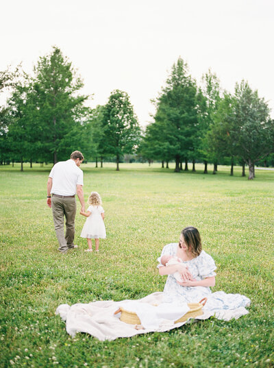 Mother sits on blanket in a green field holding newborn baby girl and father and toddler daughter walk away to explore