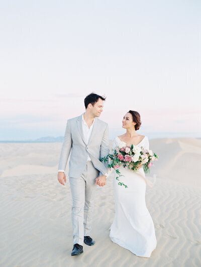 Kohnur romantic couple portrait with bride in natural subtle makeup and a sleek low bun walking on soft sand dunes at sunset.