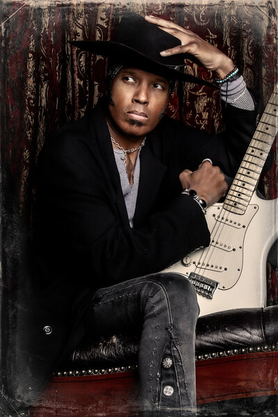 Musician photo Jeau James sitting on leather chair white guitar between legs hand on top of wide brim black hat burgundy print curtains behind him Package A