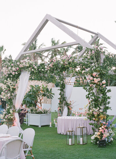 Outdoor wedding reception, round table with a white patterned table cloth, white share, and pink flowers. In the background is a white gazebo with greenery, and palm trees