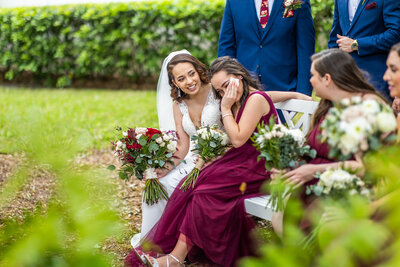 A bride and maid of honor sitting  on  a  bench with arms around each other