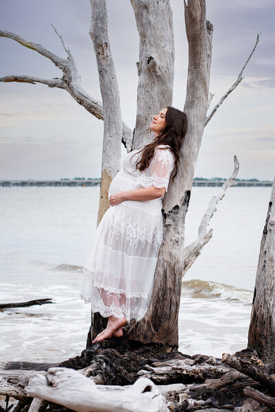 Pregnant mom in white dress leans against tree on driftwood beach in Jacksonville, FL at sunset.