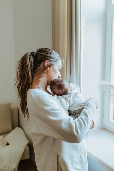 A woman holding a newborn baby and looking out a window.