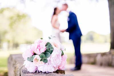 A bride and groom share a kiss in the background of an image focused on the bouquet of flowers during a wedding at Westfields Golf Club in Northern Virginia
