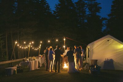 wedding guests gather around a fire pit at night with fairy lights strung up in the trees around them