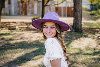 High school senior girl in purple hat posing under oak tree in New Braunfels, smiling naturally.