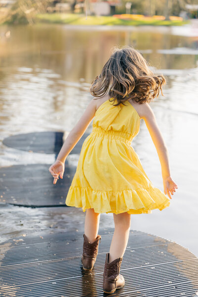A girl in a yellow spring dress and cowboy boots hops from one step to another across the pond in the Keukenhof