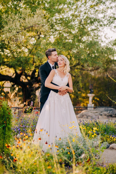 Groom embraces bride and kisses her on the forehead in a botanical gardens