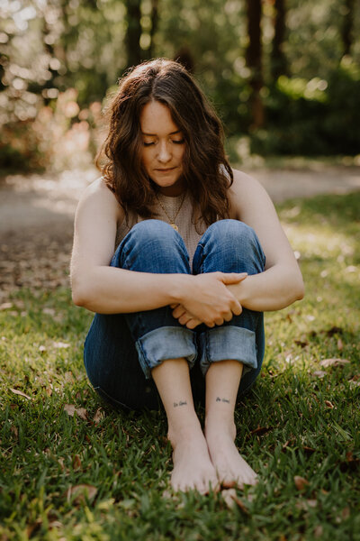 Reiki Certification Teacher, Brandi of the Intuitive Momma,  sits in grass wearing jeans looking down at shin tattoos "Be good, Do good."