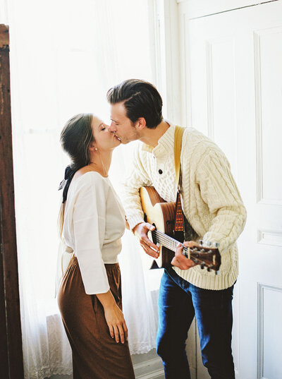 Man playing the guitar for his fiance by the window as she kisses him