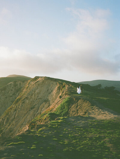 Couple in wedding attire running along  green San Francisco cliffs