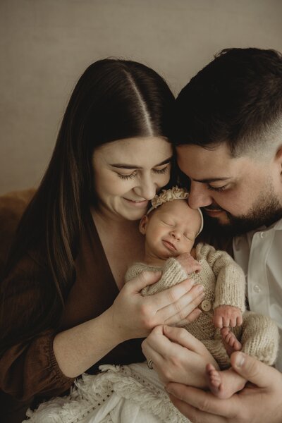 A smiling couple lovingly holding a newborn baby wrapped in a white blanket, standing close together.
