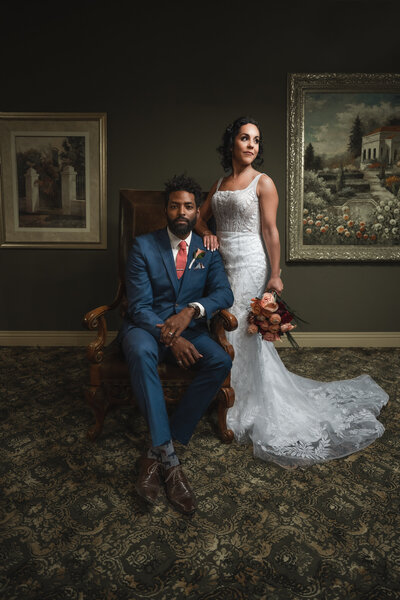 Stylish wedding portrait of a groom in a navy blue suit seated in a vintage armchair beside a bride in a lace gown holding a bouquet of red and peach flowers, posed in a room with ornate paintings and patterned carpet. Photo by Auburn Blue Photography.