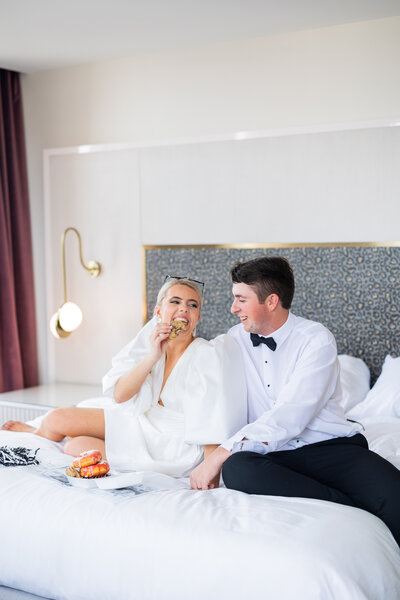 Bride and groom sitting on hotel bed eating cookies