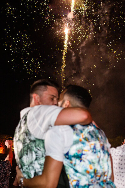 Two grooms embracing and kissing beneath golden fireworks during their Altos de Chavon Amphitheater  wedding celebration — joyful and cinematic moment captured by Asia Pimentel Photography.