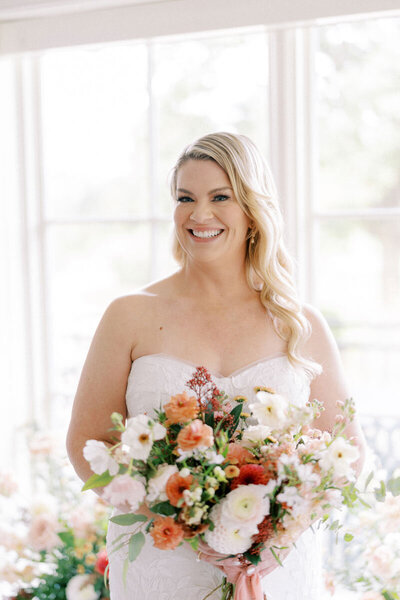 Bride in strapless gown holding vibrant bouquet, standing near window surrounded by floral arrangements