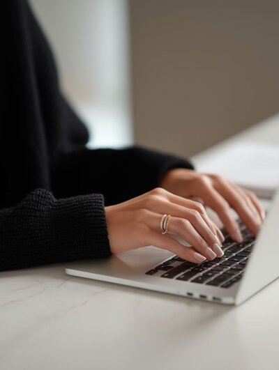Close-up of a person’s manicured hands with gold rings typing on a laptop keyboard on a white marble desk