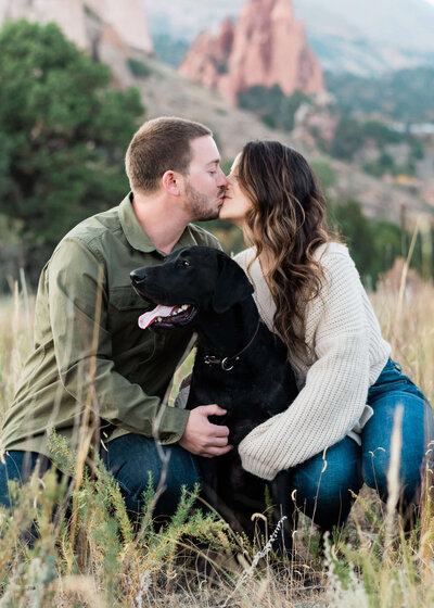 Newly engaged man and woman kiss in field with their black lab