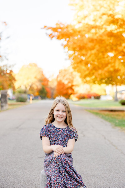 Fall photo of young girl sitting with her hands and legs crossed on a stool in the street with beautiful fall trees in the background.