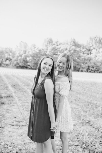 Sisters standing front to back in a field while holding hands and smiling at the camera and down at her sister.