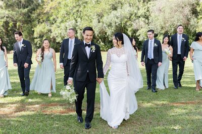 A newlywed couple holding hands with their wedding parties walking behind them 