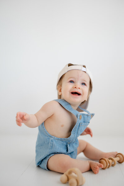 a sitting baby smiles in a white photo studio as he wears a backwards hat and denim overalls, playing with the baby toys at his feet.