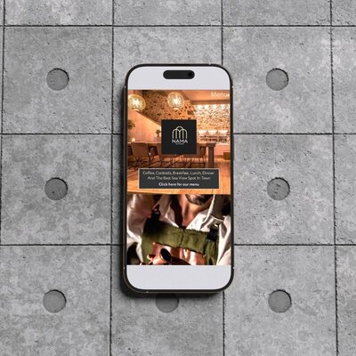 Smartphone on a tiled concrete floor showing a restaurant website with the NAMA logo, a cosy dining space, and a man in an apron holding a cocktail tool.