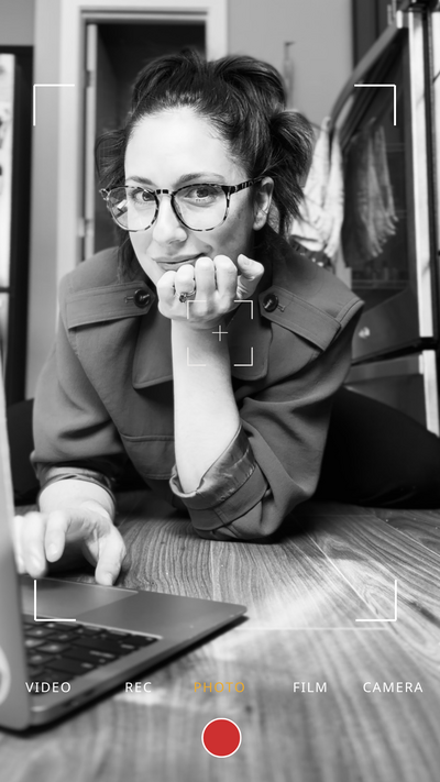 A close up shot of Hilary Rota, posed in front of her computer, with her hand under her chin, smiling and wearing glasses