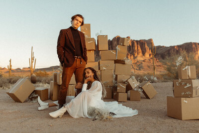 bride sitting down holding  groom's leg looking at the camera in front of boxes in the desert