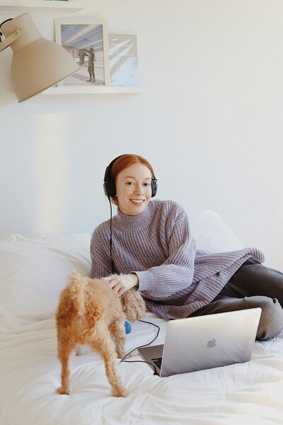 Woman relaxing on a couch with headphones on, petting her dog while using a laptop, representing a calm, supported wellness journey.