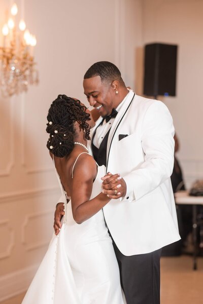 A stylish Black couple poses for an editorial wedding photo at Azaleana Manor in Orange Park, Florida. The bride sits confidently on a wooden bar ledge in a long-sleeve white mini dress with emerald heels, while the groom, in a white tuxedo jacket and black pants, leans in with a smile. Set against a warm wood-paneled wall with an arched backdrop, this modern and intimate scene captures bold elegance. Photographed by Phavy Photography, a St. Augustine wedding photographer known for editorial flair and documenting Black love stories with intention and style.