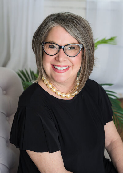 Woman sitting in beige chair wearing black shirt and glasses for a professional picture