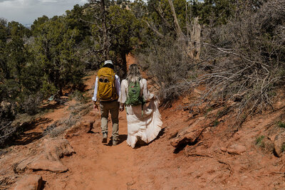 A couple hiking in Utah on their elopement day. Both are wedding their wedding attire and their backpacks. 