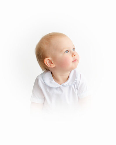 Baby in white outfit looks up softly, captured in timeless studio light with a minimal white backdrop.