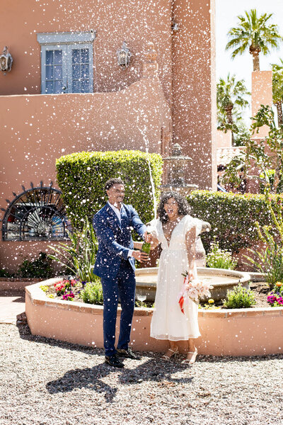 Bride and groom standing in a courtyard spraying a bottle of champagne.