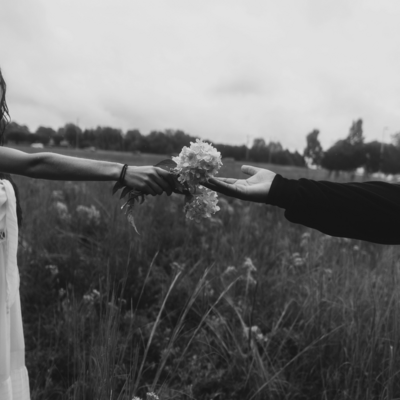 A womans arm reaches out holding white flowers from the left and a mans arm from the right reaches towards her hand
