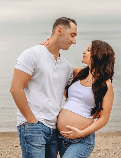 Beach maternity photography by Stamford CT photographer featuring couple in casual white outfits with ocean backdrop during golden hour session