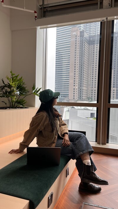 A woman in a casual outfit and green cap sits on a bench, looking out a window at tall city buildings. Nearby, a laptop and potted plants create a relaxed work atmosphere.