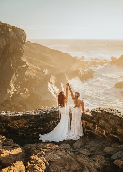 LGBTQ couple looking over the ocean at sunset in their wedding dresses