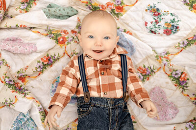 Baby boy in an orange plaid shirt and blue jean overalls is laying on a blanket smiling.