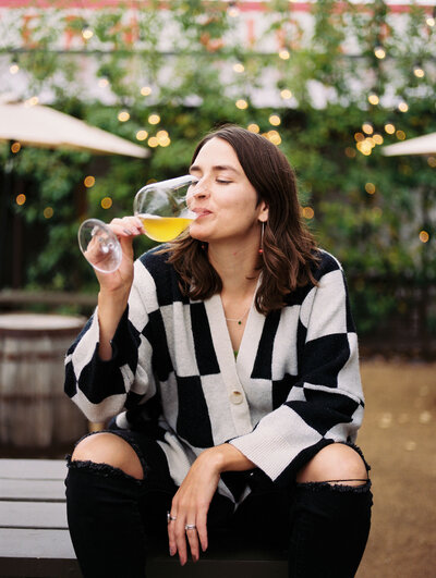 A portrait of Bright Spot founder, Emma, sipping a glass of locally made apple cider with apple trees and glowing lights in the background