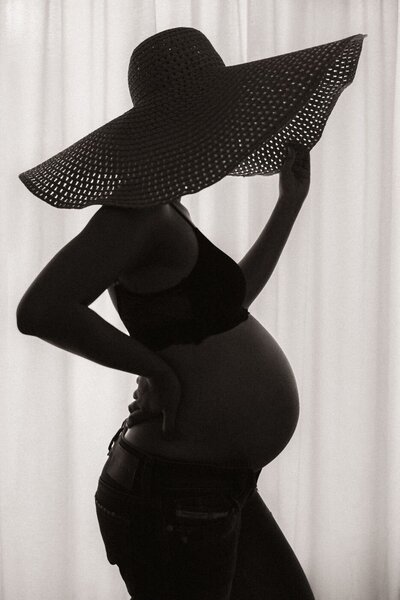 A black and white, artistic portrait of a pregnant woman's baby bump. The photo focuses on her rounded belly, with her hands gently cupping it from above and below. She is wearing a thin, sheer lace top or dress, and the soft light from the side creates a peaceful, serene atmosphere.