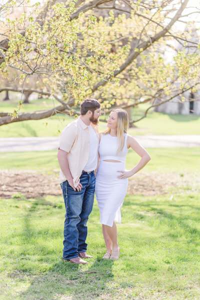A man and woman stand in front of a spring yellow tree on a green field. She is wearing a white dress and he's wearing jeans and a yellow shirt.