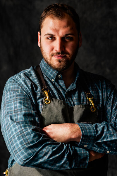 Professional male chef wearing apron posing with arms crossed in front of dark grey backdrop