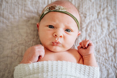 A newborn baby lying on a soft surface, wrapped in a light blanket with a green floral headband, making an expressive face with pursed lips and wide-open eyes.